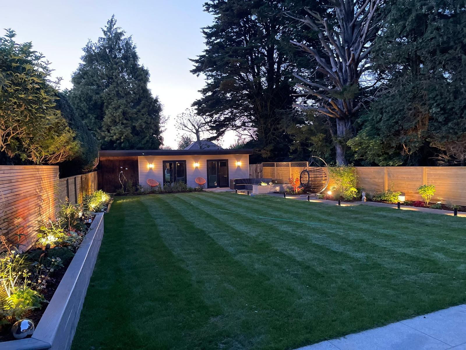 Beautifully lit garden at dusk with striped lawn, garden room and uplighting in Surrey